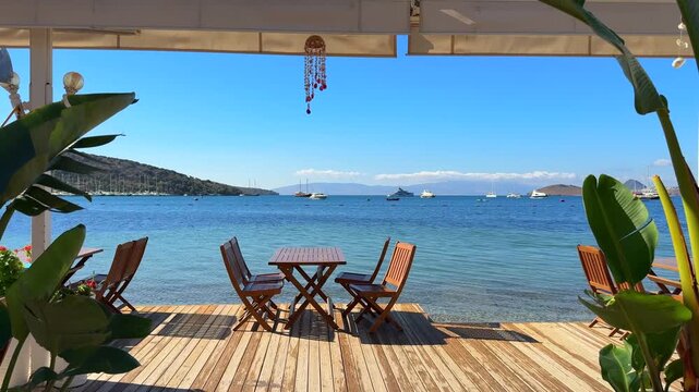 Seaside dining deck with wooden tables and chairs overlooking a beautiful turquoise bay, sailboats, and distant hills under a clear blue sky.
