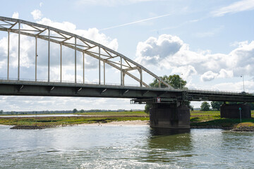 Obraz premium Steel arch bridge spanning a river against a blue, cloud-filled sky and green grassy banks.