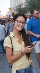 Fototapeta premium Hipster Asian woman with glasses, posing with a smile while holding a smartphone on a lively street with people in the background.