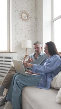 Young adult couple sitting together on sofa with laptop, discussing and planning at home. Concept of family lifestyle, partnership, online shopping, decision making and modern digital comfort