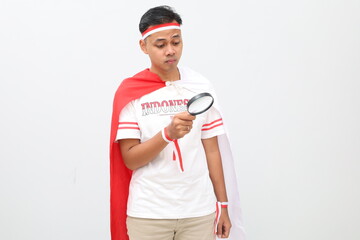 Indonesian man celebrating Indonesian independence day on August 17th while looking at something below with magnifying glass, serious expression, isolated on white background.