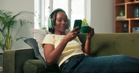 Coffee, headphones and phone with black woman on sofa in home living room for beverage break. App, scroll and music with happy person in apartment for internet search and cup of tea with laugh - Powered by Adobe