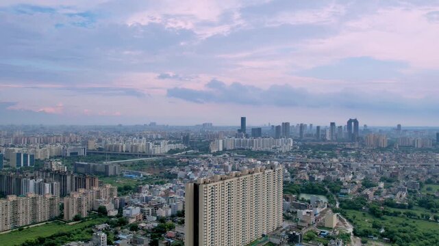 aerial drone hyperlapse flying backwards showing the multi-storey buildings in delhi, gurgaon surrounded by green fields and small houses with giant fluffy monsoon clouds flying overhead