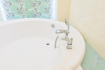 A close-up of a white bathtub with chrome fixtures. Patterned wallpaper in pastel hues can be seen behind the tub
