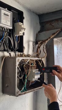 Hands of an electrician performing voltage test on exposed, messy wiring within a dirty, weathered electrical junction box. Red indicator lights illuminate on tester, showing active current.