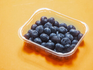 A plastic tray of blueberries sits on a bold orange background. The tray is clear and filled with fresh organic fruits ready for consumption. Healthy diet concept. Natural product.