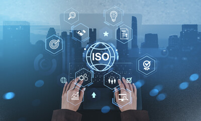 ISO certification concept with icons of quality standards, rating, and process innovation above hands typing on laptop over modern cityscape at dusk.