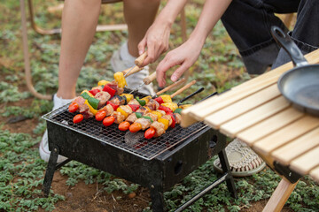 People grilling colorful meat and vegetable skewers on a portable BBQ grill during an outdoor picnic or camping, showcasing summer food, lifestyle, and social gathering atmosphere.