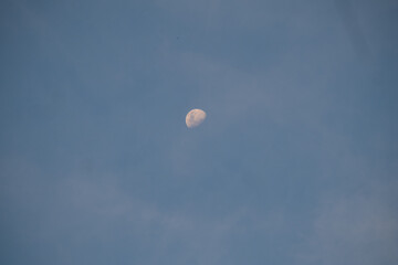 The half-Moon is visible in a vast, pale blue daytime sky with soft clouds. Minimalist and serene image offering large copy space.