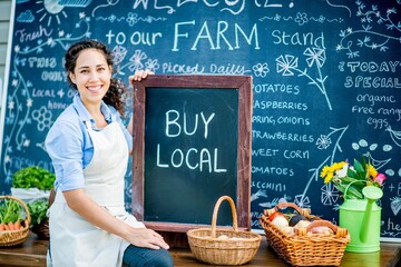Woman holding a buy local chalkboard sign in front of a farm market stand with fresh produce