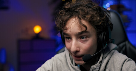 The face of a young gamer with a headset sitting in front of a computer monitor playing games child...