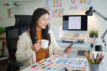 Female UX UI designer holding smartphone and coffee cup while reviewing mobile app wireframes at modern workspace.