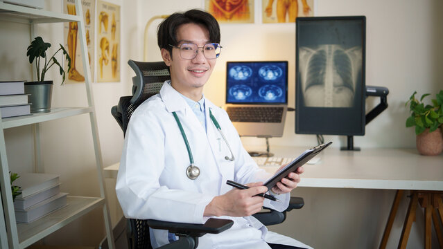 Portrait of a smiling male doctor sitting at his desk holding a clipboard in a modern medical office with X-ray images in the background.