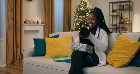 Obraz premium A young student of multi-ethnic background sits on a couch, unpacking a Christmas present. Expecting a miracle, she opens the box, pulls out a warm black hat, rejoices, and tries it on.
