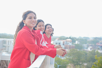 Obraz premium Three beautiful indian asian women friends standing and enjoy watching city view at balcony wearing red attire. Copy space. Female friendship.