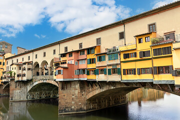 The historic Ponte Vecchio bridge in Florence, Italy. Colorful shops line the bridge, reflecting on the calm Arno river, creating a picturesque scene in bright daylight.