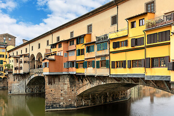 The historic Ponte Vecchio bridge in Florence, Italy. Colorful shops line the bridge, reflecting on the calm Arno river, creating a picturesque scene in bright daylight.