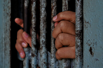 A person's hands tightly grasp the vertical, weathered metal bars of a rusty prison door or window, emphasizing feelings of confinement and despair in a stark, dimly lit setting.