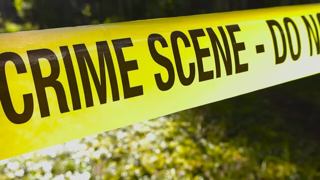 Close up video gliding over a yellow colored police line crime scene investigation forensic tape in front of a forest area, sunlight backlighting it. Shallow depth of field.