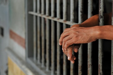 Obraz premium A person's hands tightly grasp the vertical, weathered metal bars of a rusty prison door or window, emphasizing feelings of confinement and despair in a stark, dimly lit setting.