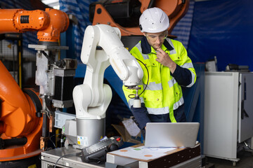 Engineer in safety gear working with industrial robotic arm, analyzing data on laptop in smart factory. Concept of automation, robotics, AI, and smart manufacturing systems.