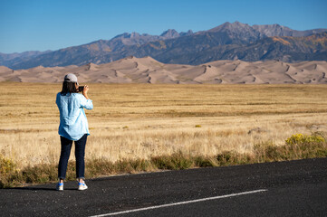 Female tourist using smartphone capturing photo of desert landscape and golden meadow in autumn against the clear blue sky at Great Sand Dunes National Park in Colorado, USA.