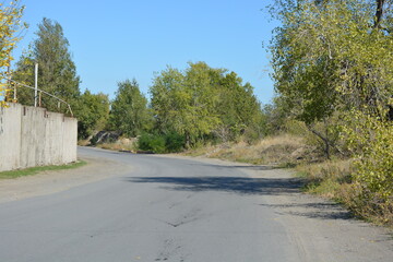 Beautiful and bright nature, a gray road with gray cedar, a fence, a curb and gray sand on the side of the road, large deciduous trees, a blue sky.