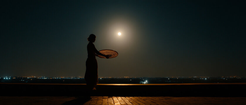Dramatic silhouette of an Indian woman on a city rooftop at night, holding a sieve (channi) while performing the ancient Karwa Chauth ritual and waiting to observe the moon