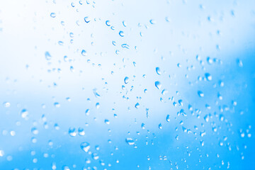Numerous clear water droplets clinging to glass surface, set against softly blurred background of bright blue and white sky. Light refracts through the drops, creating a sense of freshness and calm