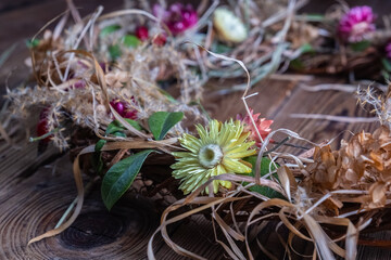 Close-up of handmade wreath with dried yellow, white, and pink flowers, green leaves, natural raffia on wooden textured background, soft lighting, horizontal orientation