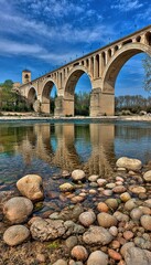 Naklejka premium Stone bridge arches over calm river with sky reflection and rocky foreground
