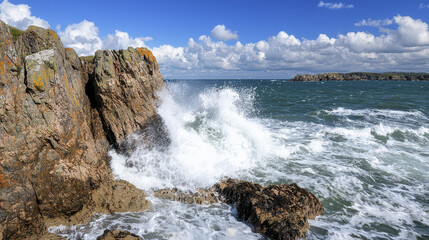 Rugged cliff with crashing waves against shore creates dramatic coastal scene under blue sky filled with clouds