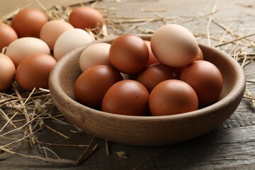 Raw chicken eggs in bowl and straw on wooden table, closeup