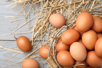 Raw chicken eggs, metal basket and straw on grey wooden table, flat lay