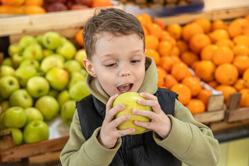 Close up happy little boy eating green apple at farmer's market with apple and tangerine counter in the background. Childhood and healthy nutrition concept. High quality photo