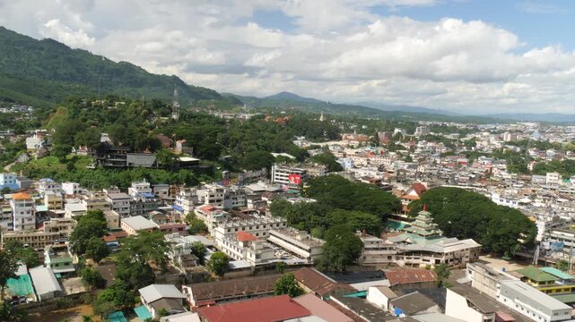 Aerial view of Tachileik town in Shan State, Myanmar (Burma), showing a mix of urban buildings, temples, and lush green hills under a cloudy sky near the Thailand border