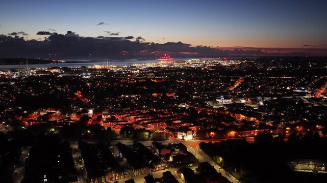 Drone descending over Liverpool at dusk, revealing warm-lit homes, red-lit cranes, and wind turbines by the water—showcasing the city’s mix of community, history, and modern energy - Powered by Adobe