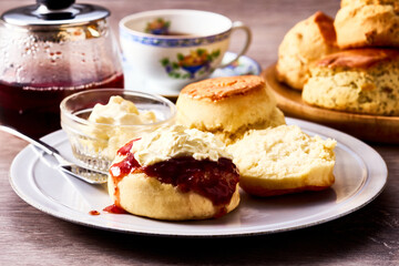 紅茶とともに楽しむアフタヌーンティーのスコーン。クロテッドクリームとジャムを添えて味わう伝統的な英国風ティータイムの一場面。ティーポットやカップと共に並べられたスコーンは、洋菓子、デザート、カフェ、休日のひとときを表現する素材写真として利用可能。