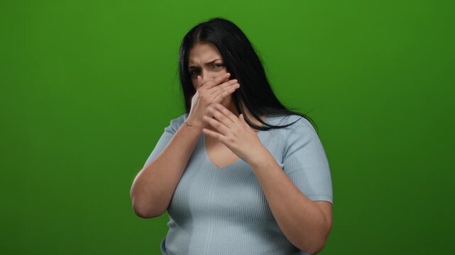 Young woman covering mouth on green background, expressing surprise or embarrassment, dressed in blue shirt, isolated and looking concerned, suitable for emotional themes.
