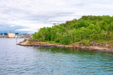  Oslofjord featuring tree covered island in foreground and distinctive Oslo city skyline in distance under cloudy sky. Beautifully captures Oslo unique blend of natural fjord beauty and urban charm