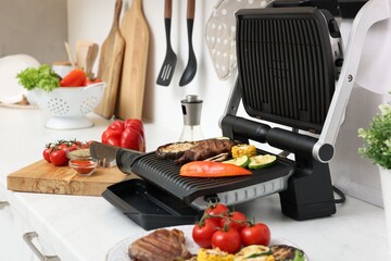 Electric grill with tasty meat, vegetables and spices on white countertop in kitchen