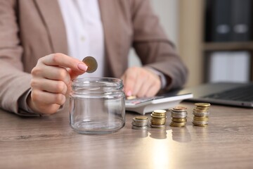 Woman putting coin into glass jar and using calculator at wooden table indoors, closeup