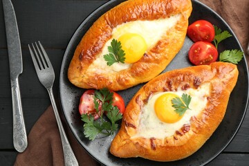 Tasty khachapuri with cheese and eggs served on black wooden table, flat lay