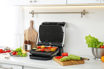 Electric grill with tasty meat and vegetables on white marble table in kitchen