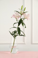 Beautiful lily flowers in glass vase on pink wooden table near white wall indoors
