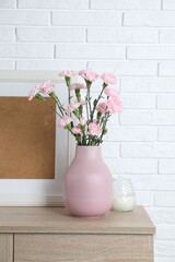Beautiful carnation flowers in vase and other decor on wooden cabinet near white brick wall indoors