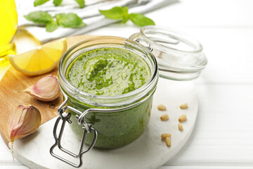 Tasty green pesto in glass jar and ingredients on white wooden table, closeup