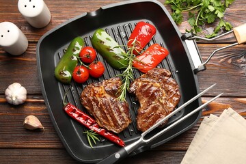 Pan with grilled beef steaks, rosemary, vegetables, shakers and carving fork on wooden table, flat lay