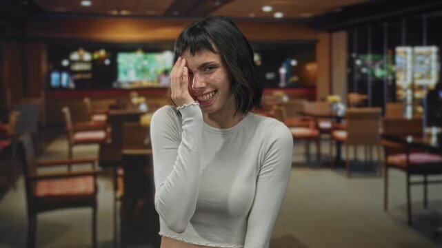 Young hispanic woman smiling broadly with hand covering face in a cozy restaurant building; embarrassment.