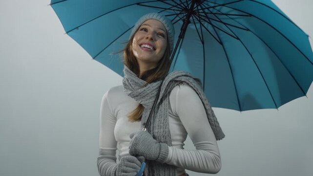 Woman wearing knit hat and scarf holding blue umbrella, smiling and looking up in gray studio backdrop with winter gloves visible; optimism.
